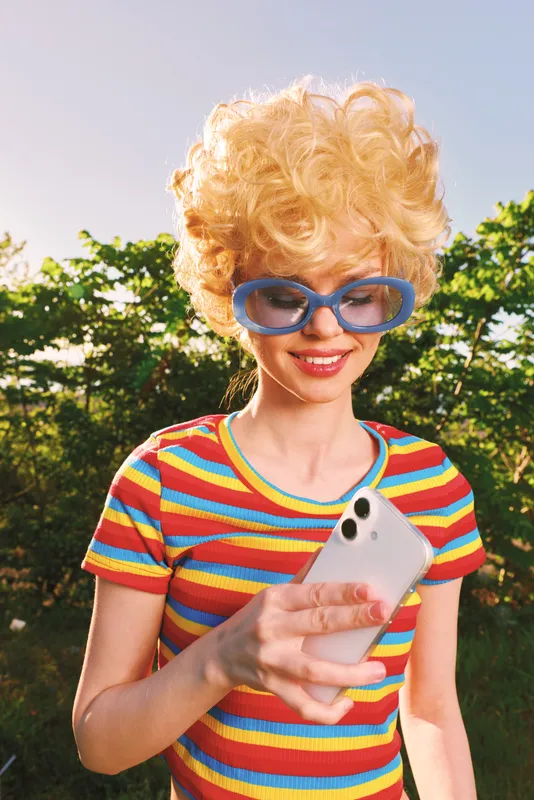 Brightly dressed woman with retro curls wearing oversized blue sunglasses, holding a smartphone outdoors. Colorful striped tee, sunny garden backdrop, playful vibe, casual lifestyle, bold colors.