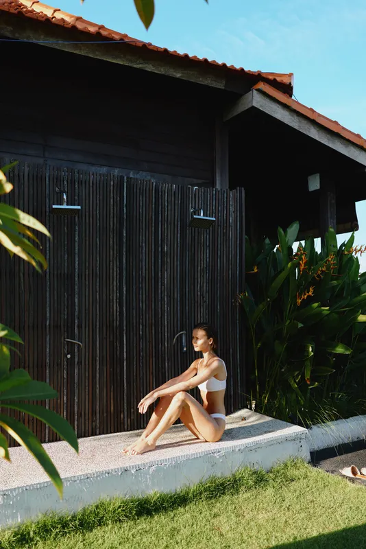 Woman in a bikini sits on a concrete ledge beside a wooden wall, tropical plants surrounding. Outdoor sunlit scene conveys relaxation, leisure, wellness, and vacation mood with warm natural lighting.