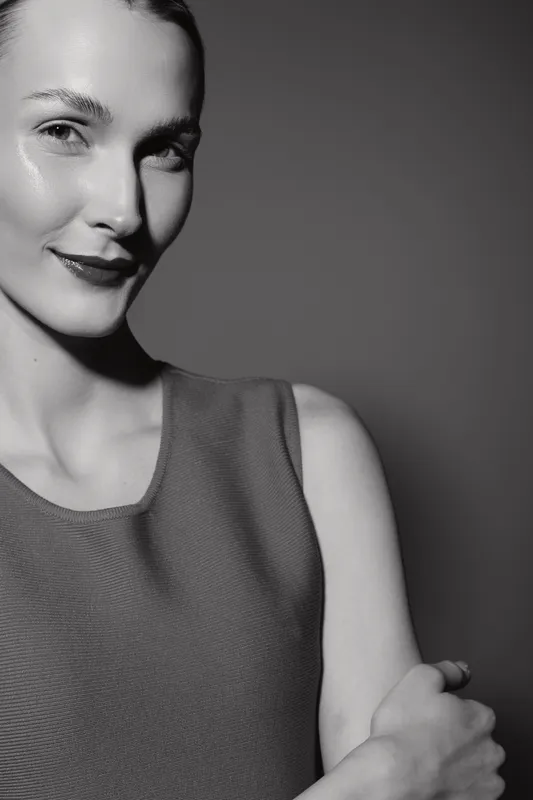 Portrait of a smiling white woman with light skin and slicked hair wearing sleeveless top in studio with dark background.