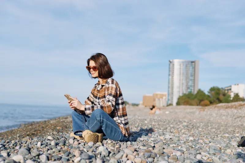Woman on a pebble beach using a smartphone, casual plaid shirt and jeans, enjoying outdoor relaxation by the sea with distant city buildings and blue sky.