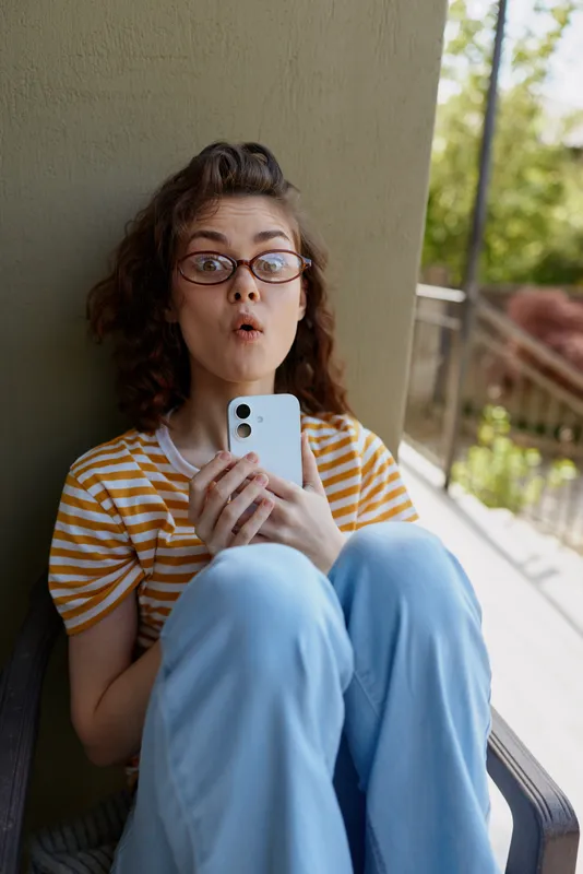 Surprised woman with curly hair wearing a striped shirt and glasses sits on a balcony, holding a smartphone. Casual home moment, playful mood, candid lifestyle portrait.