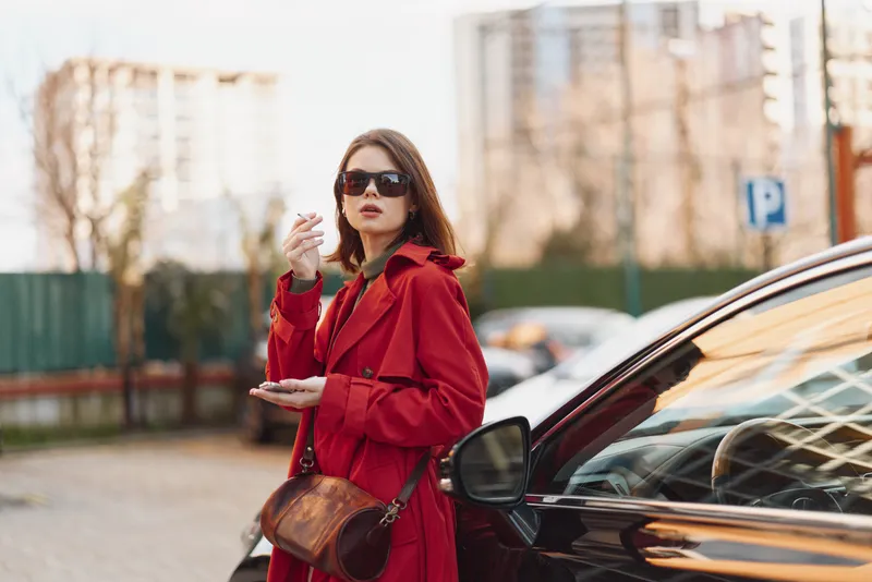 One woman in a red trench coat stands beside a sleek car in a city parking lot, wearing sunglasses and a brown crossbody bag, gazing confidently on a sunny day.