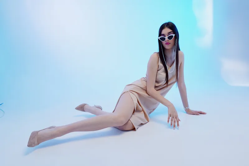Fashionable woman in a beige dress poses on the floor wearing white sunglasses. Studio shot with soft lighting on a light blue background. Concept of beauty, fashion, and elegance.