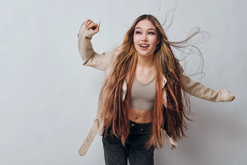 Young woman with long hair is smiling and dancing wearing casual clothes and jacket on grey background. Female model with joyful expression and dynamic pose for lifestyle and fashion.