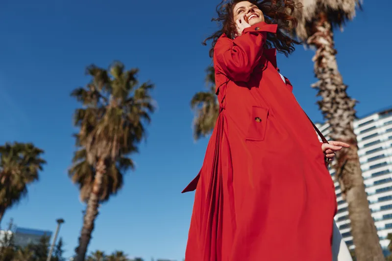Young woman in a red coat smiles joyfully while walking outdoors on a sunny day with palm trees and modern buildings in the background.