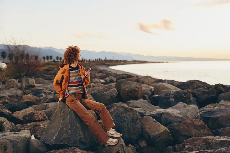 Woman lifestyle outdoor sunset beach rocks casual sitting calm nature, curly haired woman sits on beach rocks at golden hour, wearing casual jacket and jeans, warm light, ocean horizon