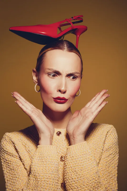 A confused woman with red lipstick and gold earrings balances a red high heel shoe on her head. She wears a textured beige jacket and poses against a warm brown background.