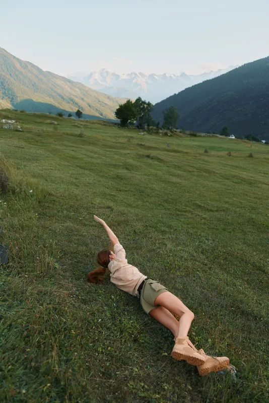 Young woman relaxing on a hillside meadow with mountains in the background, stretching her arm, casual outdoor lifestyle scene suitable for stock, bright atmosphere, natural light
