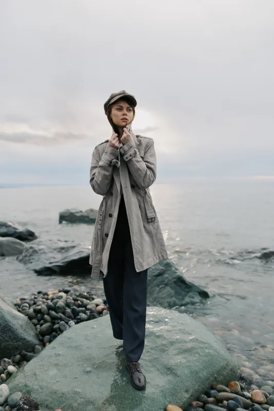Young woman in trench coat and cap stands on rocky shore near calm sea under cloudy sky. Concept of autumn fashion, outdoor lifestyle, and thoughtful mood by the water.