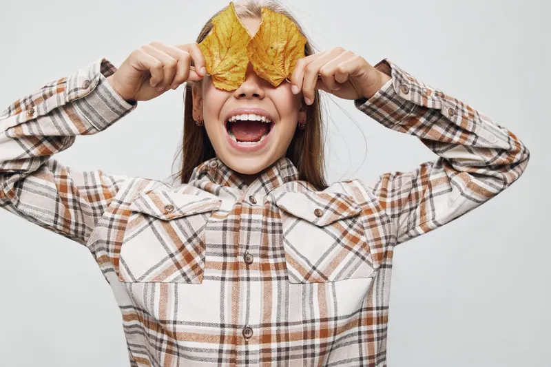 Child smiling and playing with autumn leaves covering eyes, wearing plaid shirt in studio. Girl with open mouth expression, seasonal fun and casual outfit on white background.