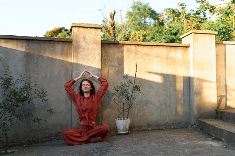 Young woman in a red dress sits cross legged on a rooftop, raising her arms to form a heart above her head, conveying calm, joy, and playful serenity