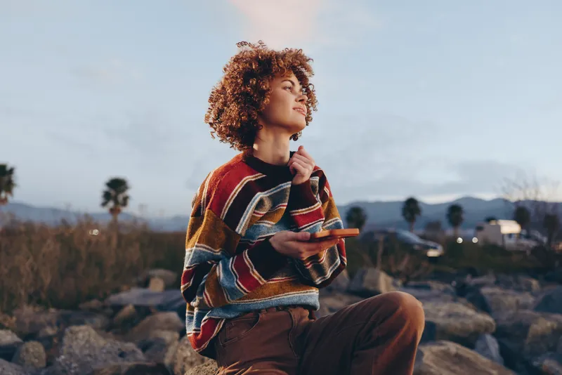 Woman lifestyle outdoor portrait, young curly haired woman in patterned sweater sits on rocky shore, gazing into distance with calm smile, hands near chin, sunset sky, soft light