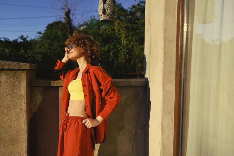 Cheerful woman outdoors, wearing orange jacket and yellow top, standing on a sunlit balcony, relaxed pose, soft breeze, vibrant colors, natural backdrop, urban greenery