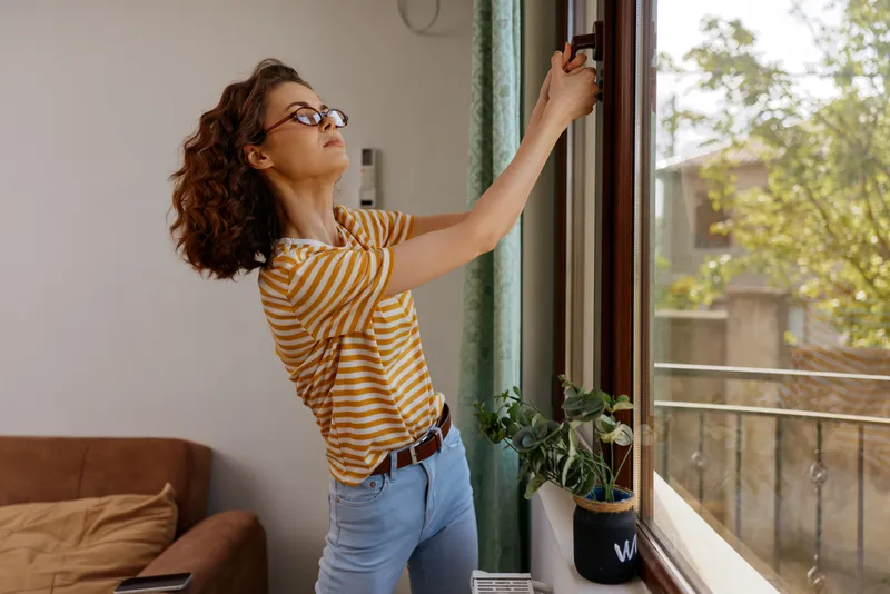 Cheerful woman at home, standing by an open window, wearing a striped shirt, relaxed posture, sunlight on her face, soft indoor light, greenery outside, candid lifestyle scene