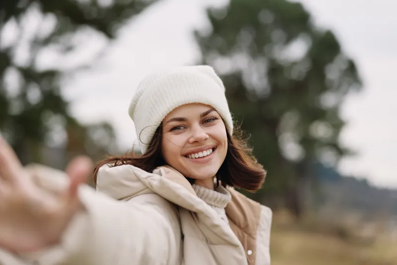 Smiling woman taking a selfie outdoors in winter, wearing a warm jacket and beanie, enjoying a bright moment in nature with soft bokeh background and relaxed fashion vibes.