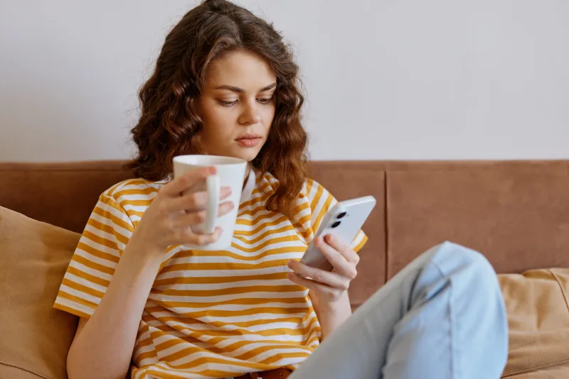 A woman sits on a sofa at home, sipping from a mug while scrolling on her smartphone, a calm, everyday moment. Concept: relaxed lifestyle, domestic routine.
