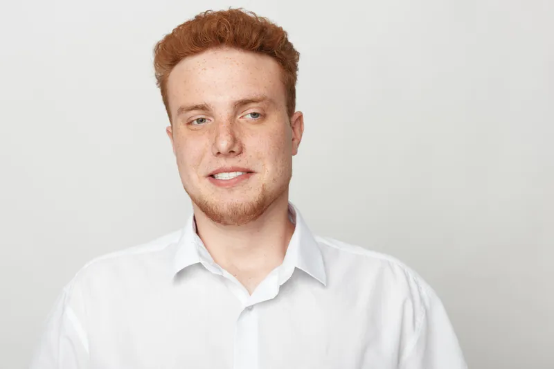 Portrait of a young redhead man with freckles wearing a white shirt against a plain light background. Casual style, natural expression, and soft lighting for a clean look.