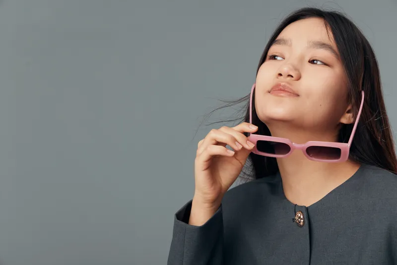 Young woman holding pink sunglasses and looking away thoughtfully in studio. Female portrait with long hair wearing dark jacket on gray background for fashion style and beauty.