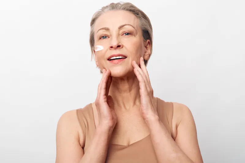 Mature woman with smooth skin applies cream on her face in soft natural light. Concept of skincare, anti aging, beauty routine, wellness, and self care for healthy skin.