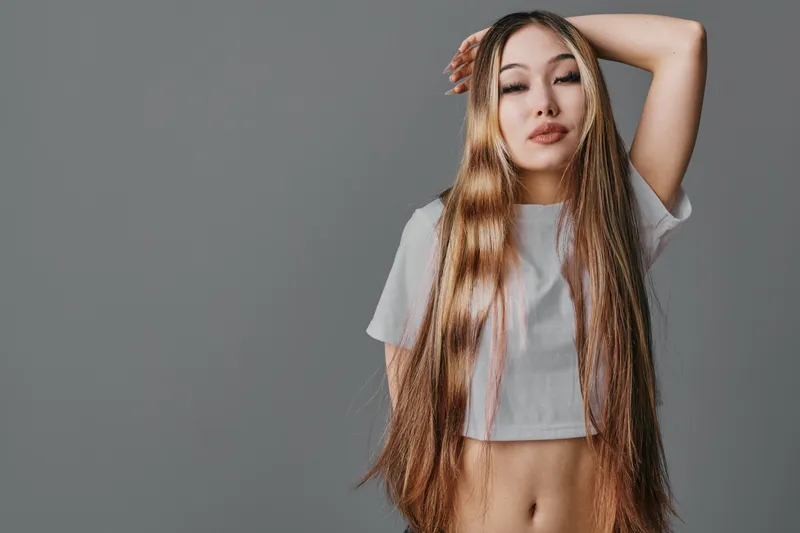 Woman with long natural hair and hand raised posing in white crop top against grey background. Beauty, fashion, hairstyle and confidence with casual clothing and relaxed posture.