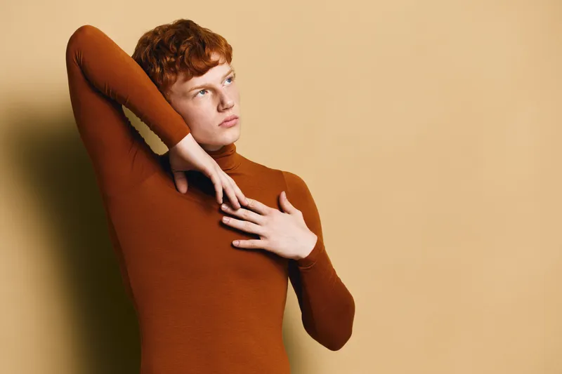 Young redhead man with curly hair wearing brown turtleneck posing thoughtful with arm raised against beige background in fashion portrait studio shoot.