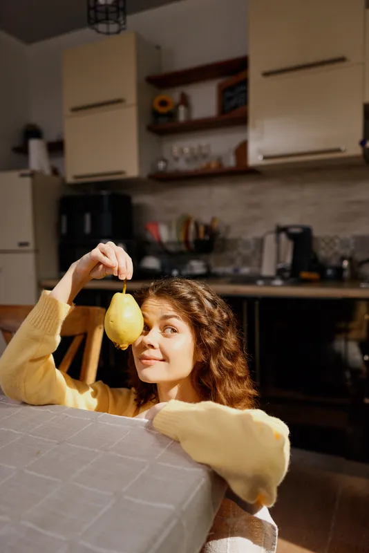 Cheerful woman indoors at a kitchen table, holding a lemon above her head, warm light, casual pose, cozy home setting, soft colors, candid lifestyle moment