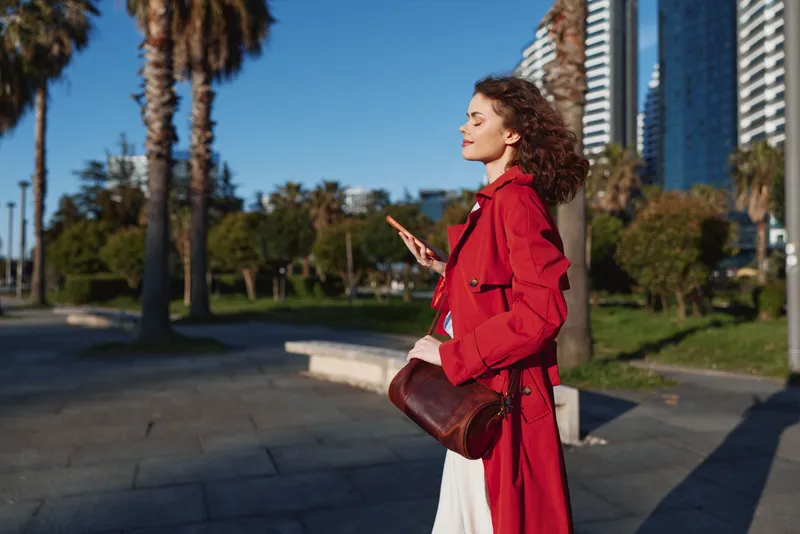 Woman in a red trench coat walks along a sunny urban promenade, phone in hand, brown crossbody bag, enjoying a calm city moment with skyscrapers behind.