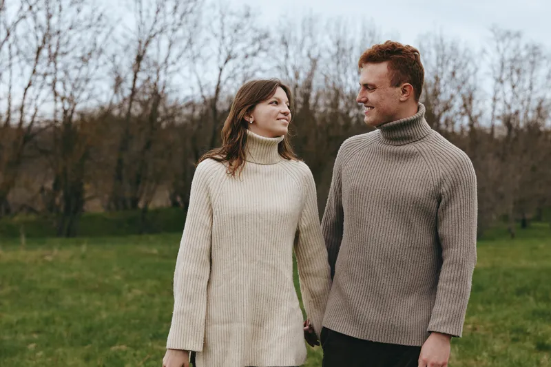 Happy couple strolls in a park wearing chunky knit sweaters, holding hands, smiling at each other, an outdoor lifestyle moment with autumn tones and bare trees in the background, warm romance.