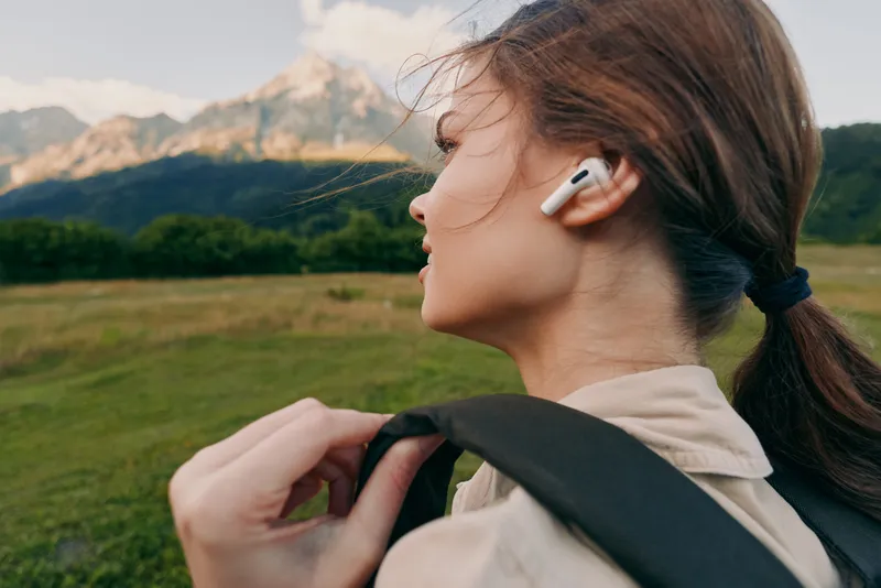 A woman with a backpack walks outdoors wearing wireless earbuds, enjoying fresh air in a wide valley with distant mountains, capturing a relaxed, everyday lifestyle moment.