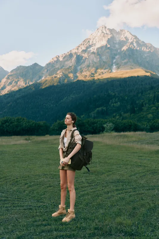 A woman hiking in a grassy meadow with tall mountains in the background, wearing boots and a backpack, enjoying nature and a calm outdoor lifestyle