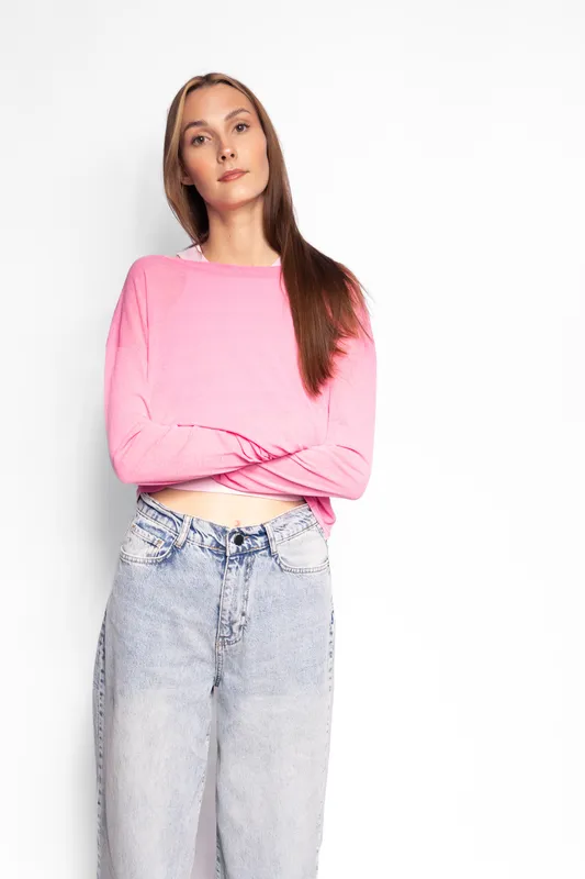 Young woman with long brown hair wearing a pink sweater and blue jeans stands with crossed arms against white background. Casual style and confident expression concept.