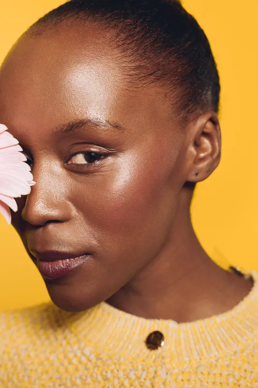 Close-up portrait of a black woman with dark skin and natural makeup holding a pink flower near her face. Yellow background and soft lighting highlight her glowing skin and calm expression.