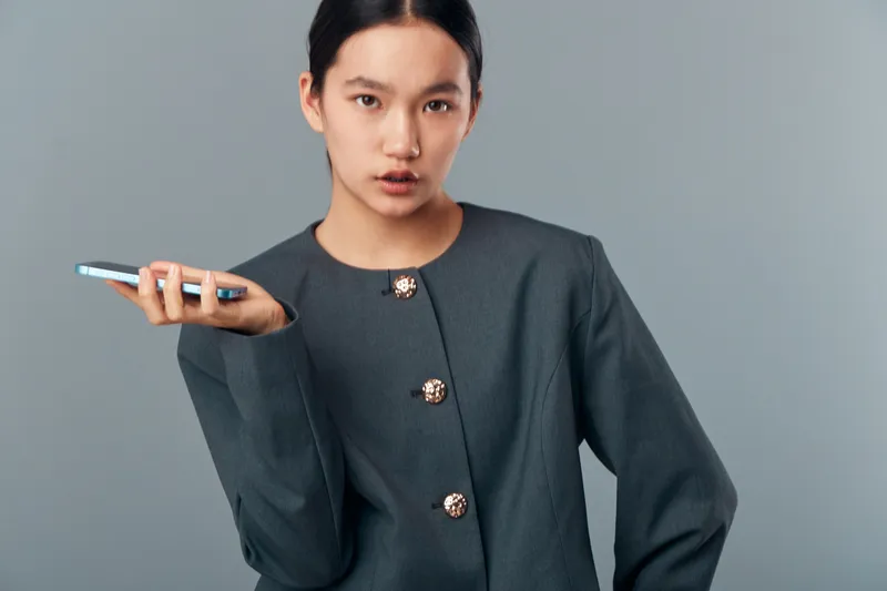 Young woman with dark hair wearing gray jacket, holding phone in one hand, looking at camera with confident expression on neutral gray background in studio lighting.