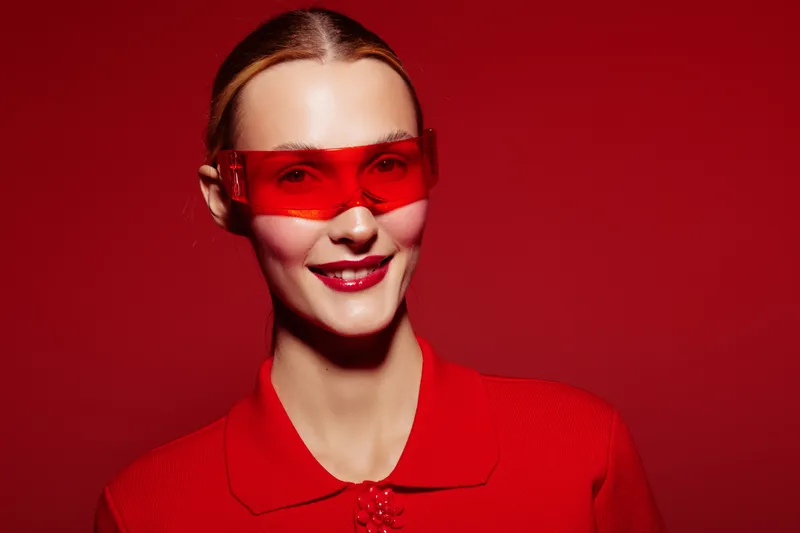 Smiling woman with red sunglasses and red outfit poses against red background. Studio portrait with bright lighting and confident expression. Concept of fashion, style, and bold colors.