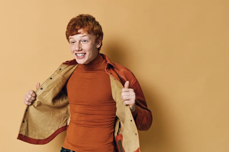Young man with red hair wearing orange sweater and jacket showing excitement and smiling against plain background. Studio portrait with casual fashion and positive mood.