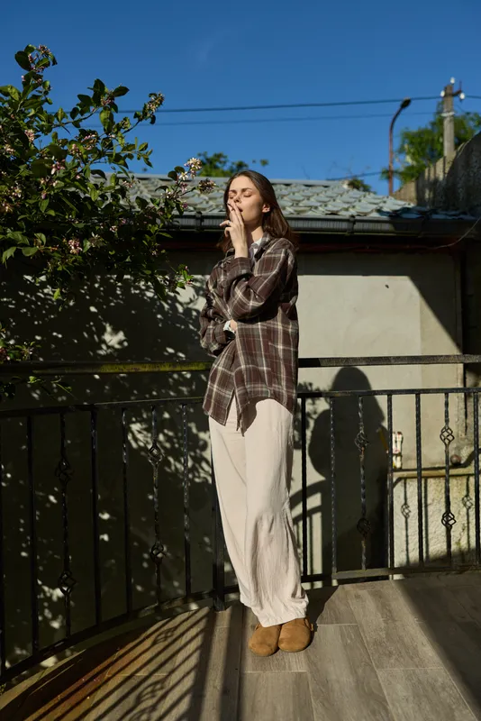One person on a sunlit balcony wears a plaid shirt and white pants, standing relaxed as they blow a soft kiss to the breeze
