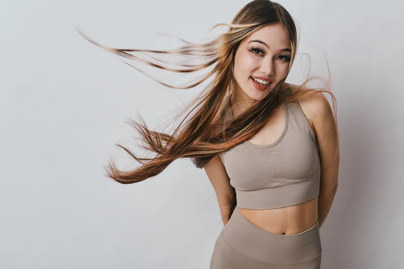 Young woman with long flowing hair smiling and posing in beige sportswear against white background for beauty, fitness and healthy lifestyle concept.
