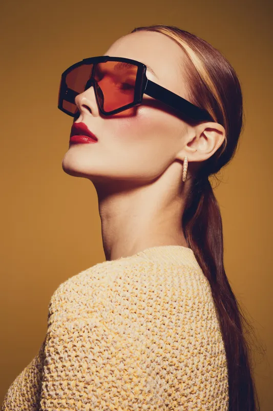 Stylish woman with sleek hair wears oversized sunglasses and red lipstick. She poses confidently against a warm brown background. Concept of fashion, beauty, and modern style.