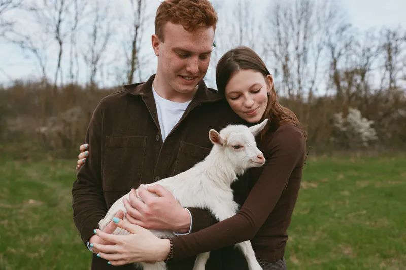 Couple outdoor goat kid moment, warm embrace, calm field, gentle breeze, soft colors, natural textures, authentic lifestyle, rural backdrop, peaceful companionship