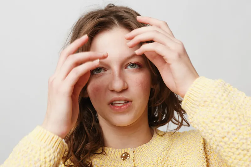 Young woman with curly hair wearing yellow sweater touches her face with hands, looking at camera on white background. Concept of natural beauty, skincare, and casual style.