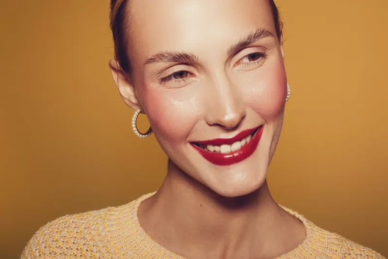 Smiling woman with red lipstick and hoop earrings looks away on yellow background. Close-up portrait with natural skin texture and warm lighting. Concept of beauty, makeup, and happiness.