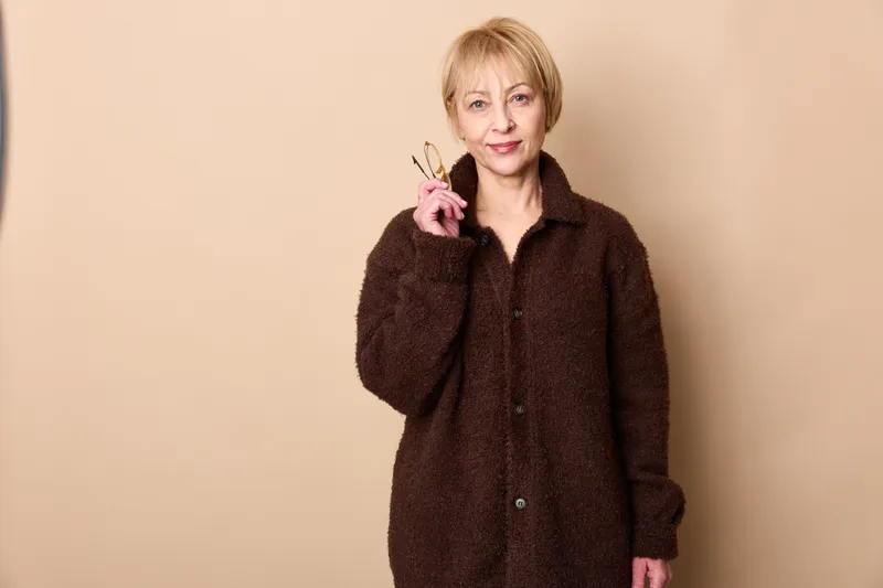 Mature woman in studio portrait wearing brown coat, raised finger gesture, smiling softly, beige background, standing relaxed, calm confident mood indoors