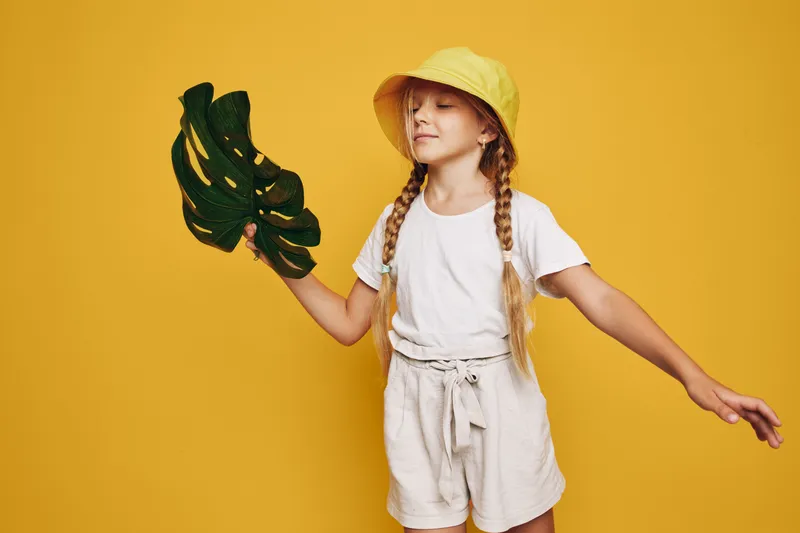 Young girl in yellow hat holds large green leaf with eyes closed, standing against bright yellow background. Concept of nature, summer, and childhood.