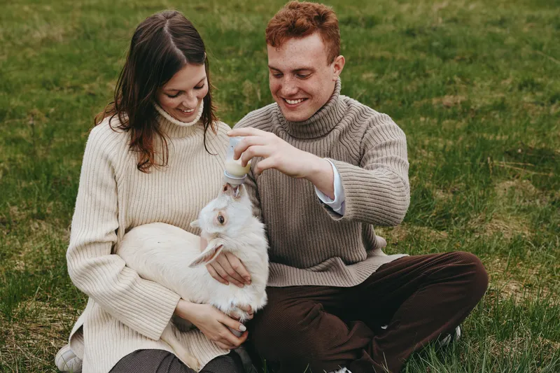 Happy couple outdoors with a white goat kid, feeding it from a bottle, cozy sweaters, relaxed mood, green field background, affectionate interaction, and a simple lifestyle moment in nature
