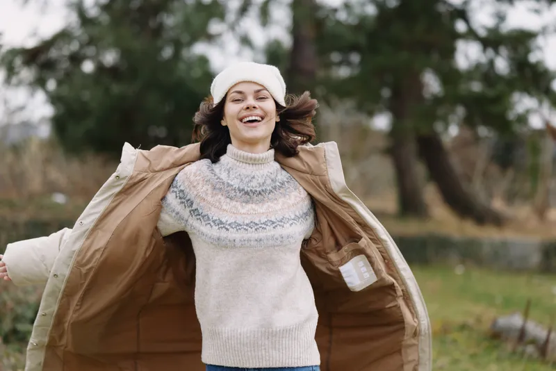 Woman outdoors wearing a knitted sweater and beanie, joyful and energetic as she swings open a warm coat, capturing movement against a park backdrop with trees and soft light