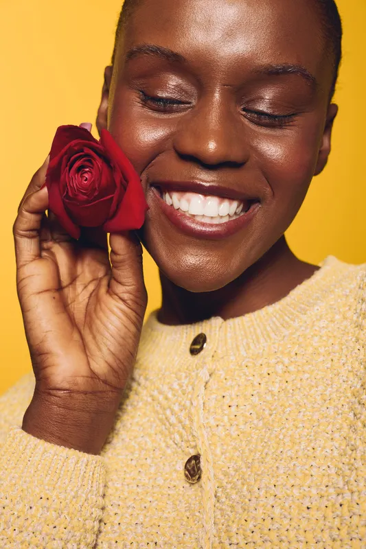 Smiling dark skinned African woman holding red rose near face with closed eyes. Yellow background and cozy knitted sweater. Concept of beauty, happiness, nature, and self-love.