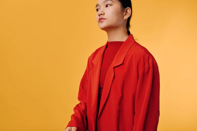 Young woman in red jacket standing against yellow backdrop looking away thoughtful and confident with casual hairstyle and minimal makeup for fashion or portrait concept.