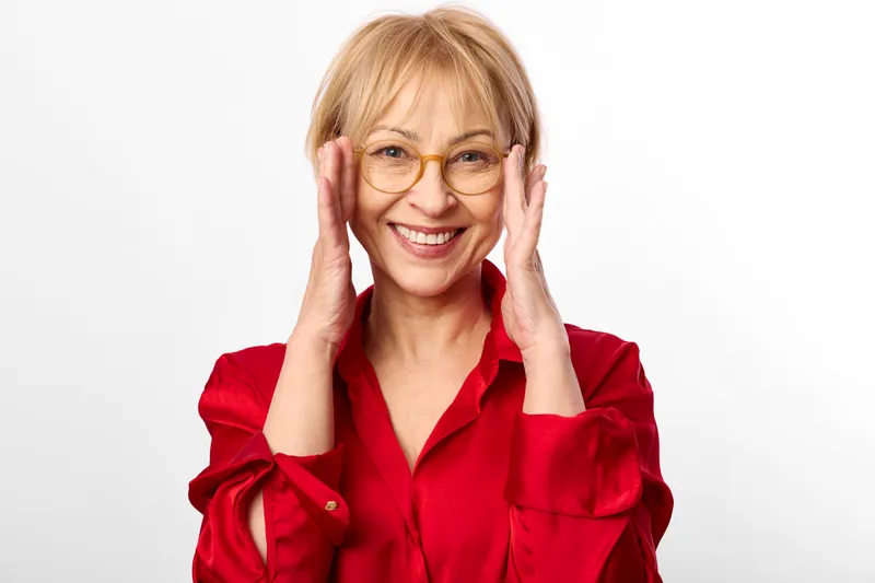 Smiling woman in red blouse and glasses, hands on cheeks, studio portrait against white background, cheerful emotion, soft even lighting, close up framing, neutral tone