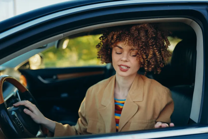 Curly haired woman driver in car, relaxed and focused, wearing a beige blazer and striped top, interior shot. Lifestyle, travel, independence, retro color, film vibe, warm mood.