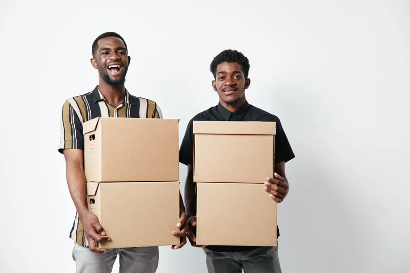 Two young black men holding cardboard boxes for moving or delivery, smiling and standing against white background. African ethnicity, dark skin, casual clothing and teamwork concept.
